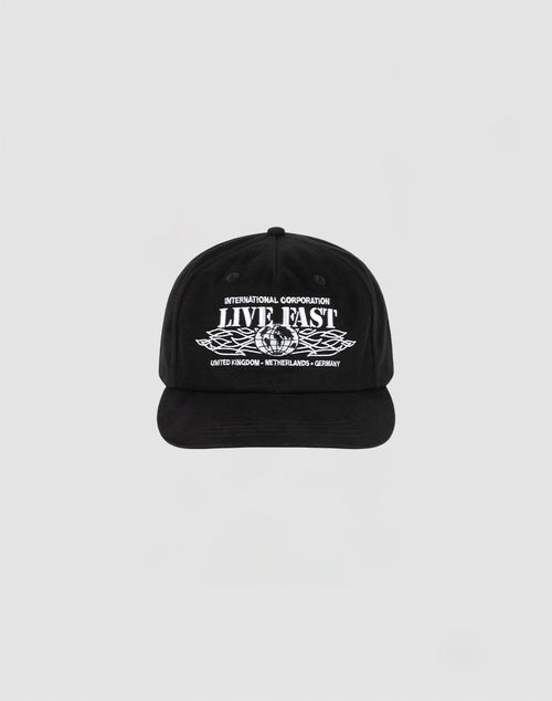 The Live Fast Corporation Cap by LIVE FAST DIE YOUNG is a black, 100% cotton unstructured 5-panel hat with multi-logo white text and graphics reading INTERNATIONAL CORPORATION LIVE FAST above stylized wings and UNITED KINGDOM - NETHERLANDS - GERMANY below.