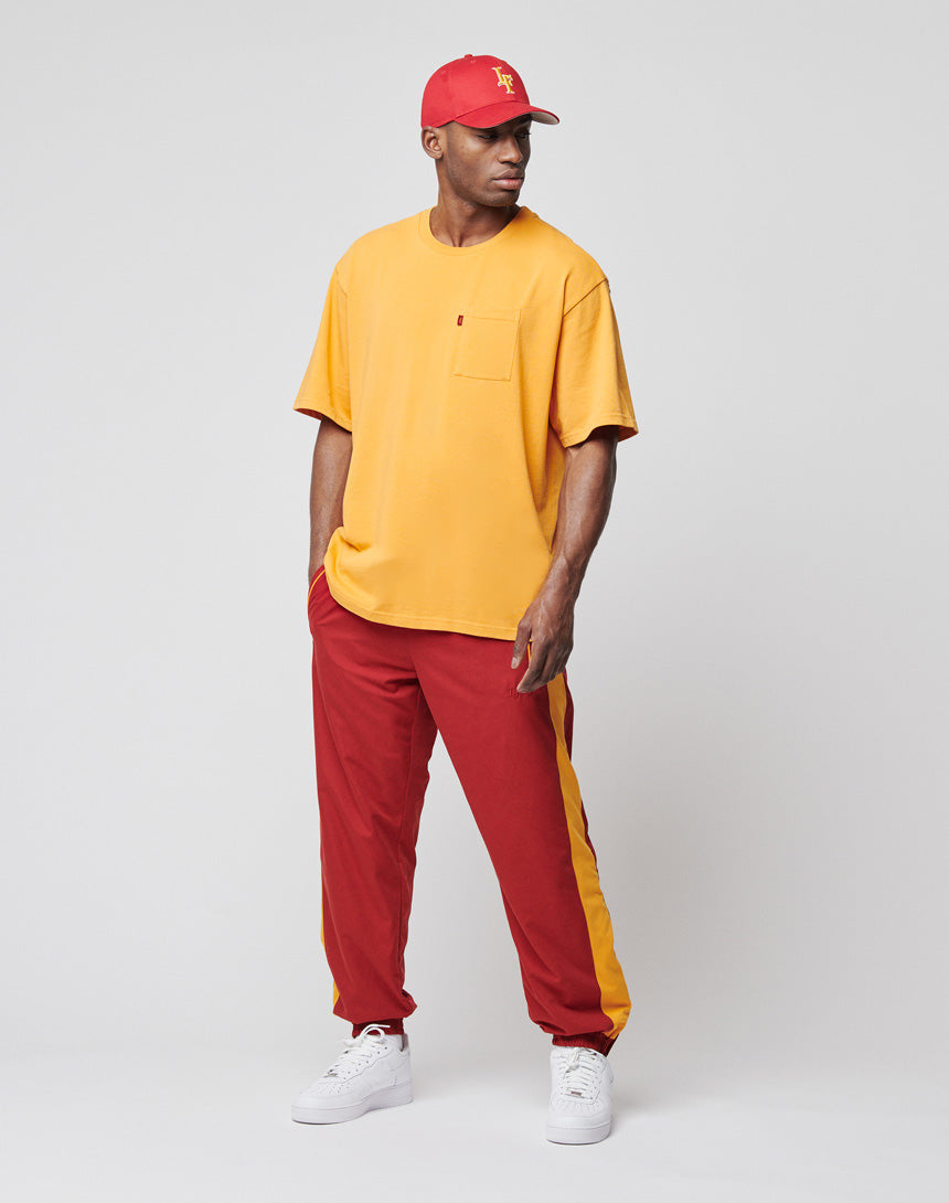 A man wearing an oversize LIVE FAST DIE YOUNG LFDY Tube Pocket Tee in yellow, with a red baseball cap, red track pants featuring yellow side stripes, and white sneakers stands against a plain light gray background.