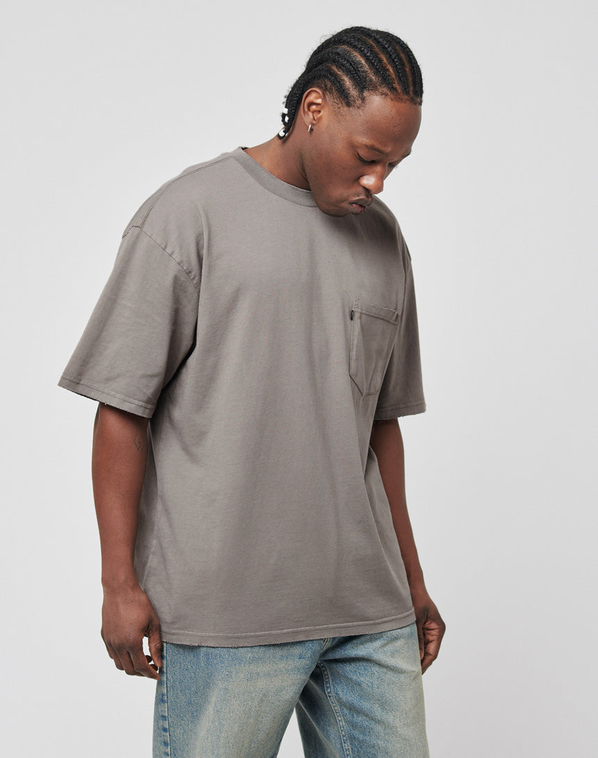 A person with braided hair looks down while wearing the LFDY Destroyed Pocket Tee in an oversize fit and light blue jeans, standing against a plain light background.
