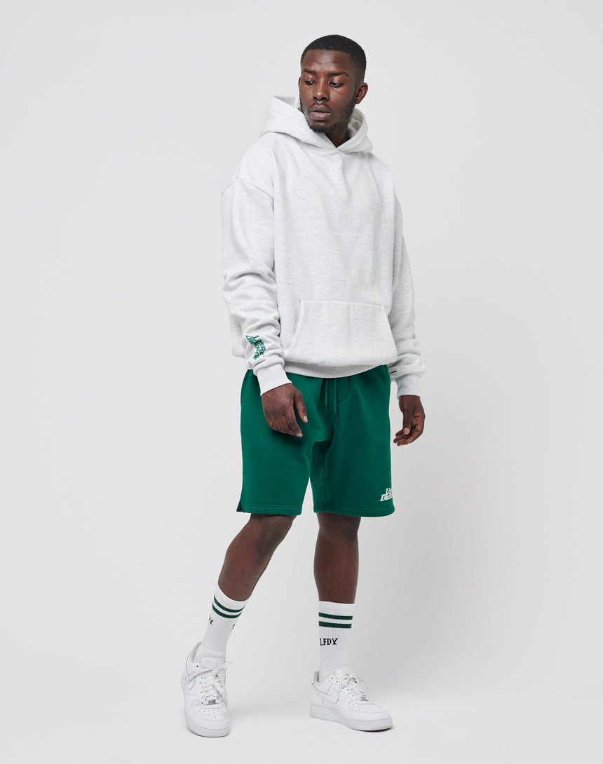 A man stands against a plain white background wearing a light gray LIVE FAST DIE YOUNG Basic Wrist Hooded, green shorts, white crew socks with black stripes, and white sneakers. He looks slightly left with one hand by his side and the other slightly raised.