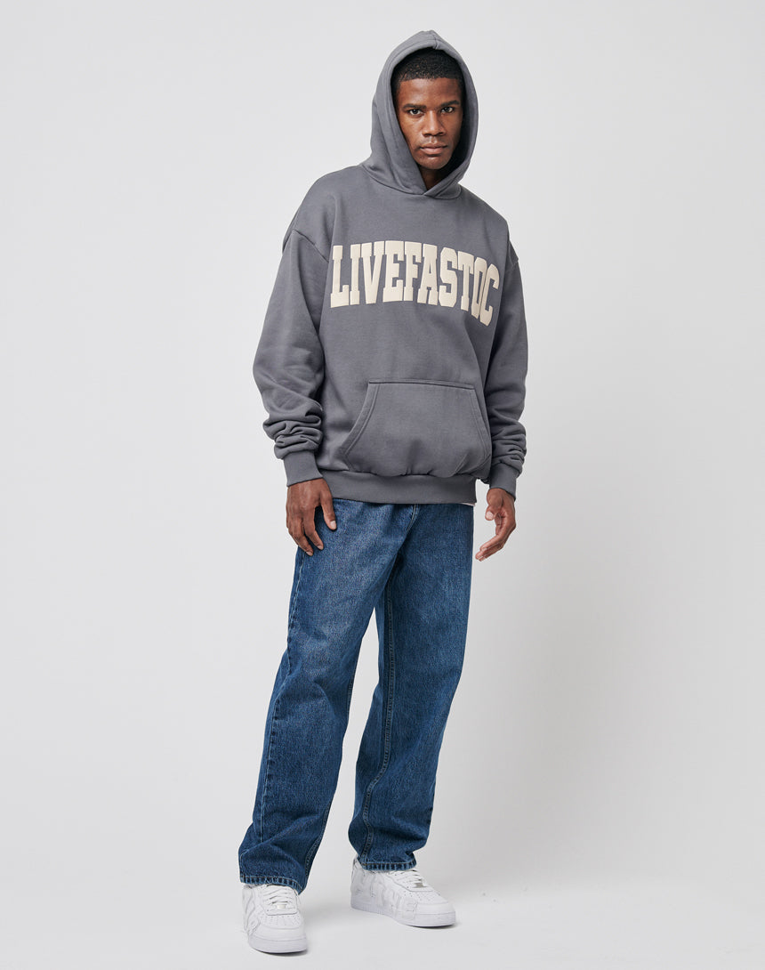 A man stands against a plain background wearing a gray hoodie with "LIVEFASTIC" in large white letters, LFDY Wide Denim jeans (100% cotton), and white sneakers.
