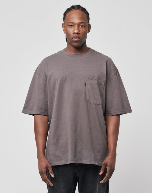 A man with braids wears the LFDY Destroyed Pocket Tee—an oversize, faded gray T-shirt with a chest pocket—paired with black jeans, standing against a plain light gray background.