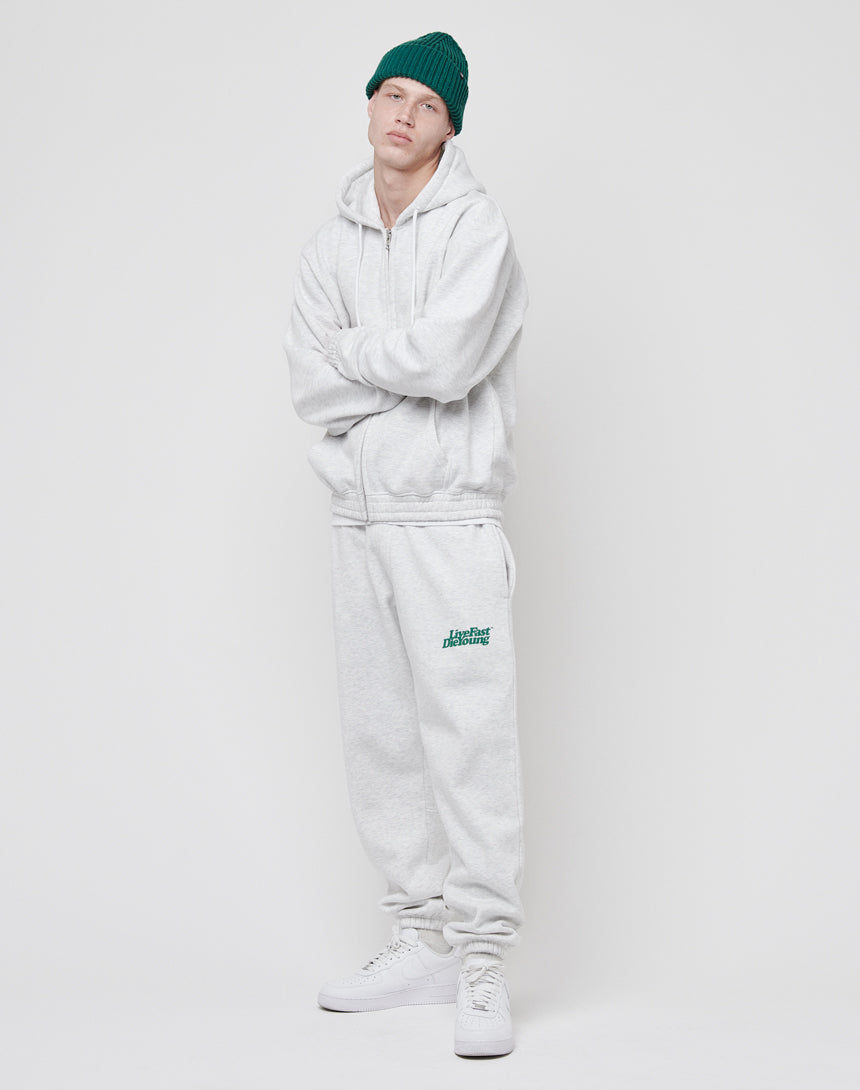 Wearing the LIVE FAST DIE YOUNG Basic Hooded Zip in light gray with matching sweatpants, white sneakers, and a green beanie, a person stands against a white background looking at the camera, arms loosely crossed.