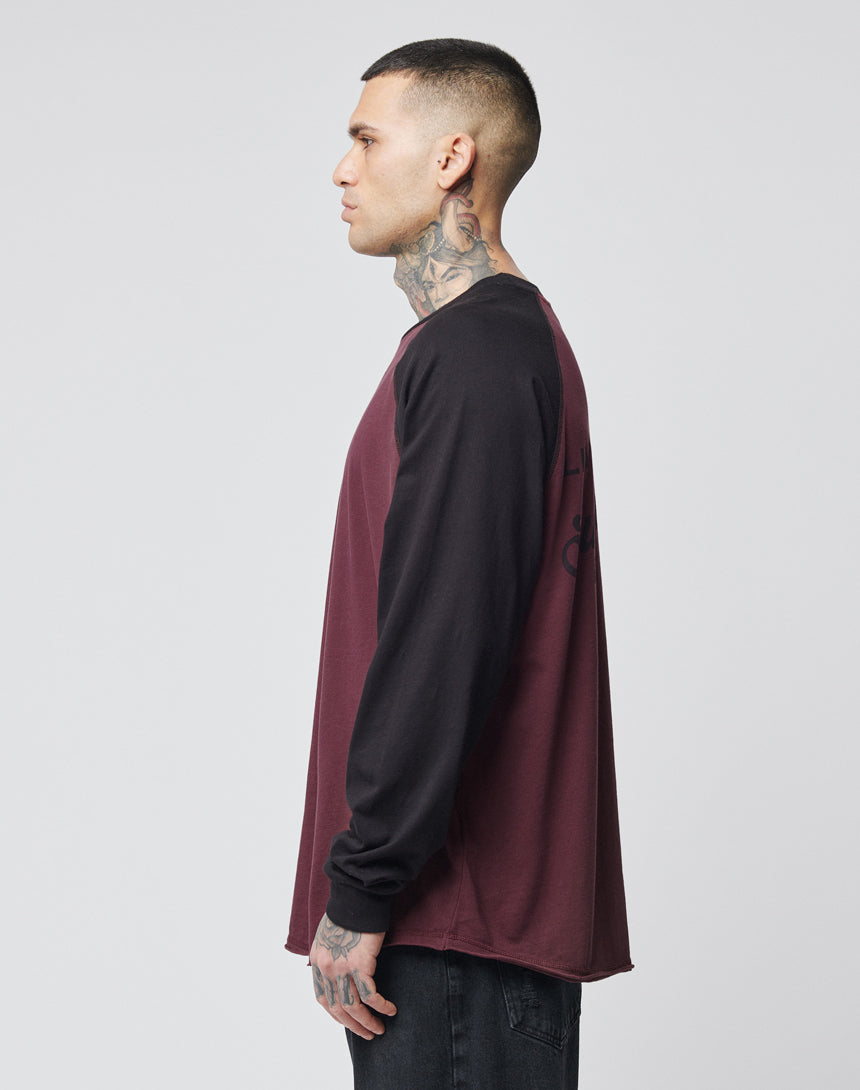 A person with neck and hand tattoos stands in profile, wearing the LIVE FAST DIE YOUNG Survival Longsleeve—an oversize fit with burgundy panels, black sleeves, and contrast cuffs—paired with dark pants against a plain light background.