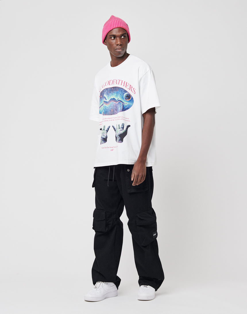 A man wears a pink beanie, loose black cargo pants, white sneakers, and the LIVE FAST DIE YOUNG YNG Godfathers Tee in Oversize Fit with colorful graphics and Vintage Effekt, standing against a plain white background.