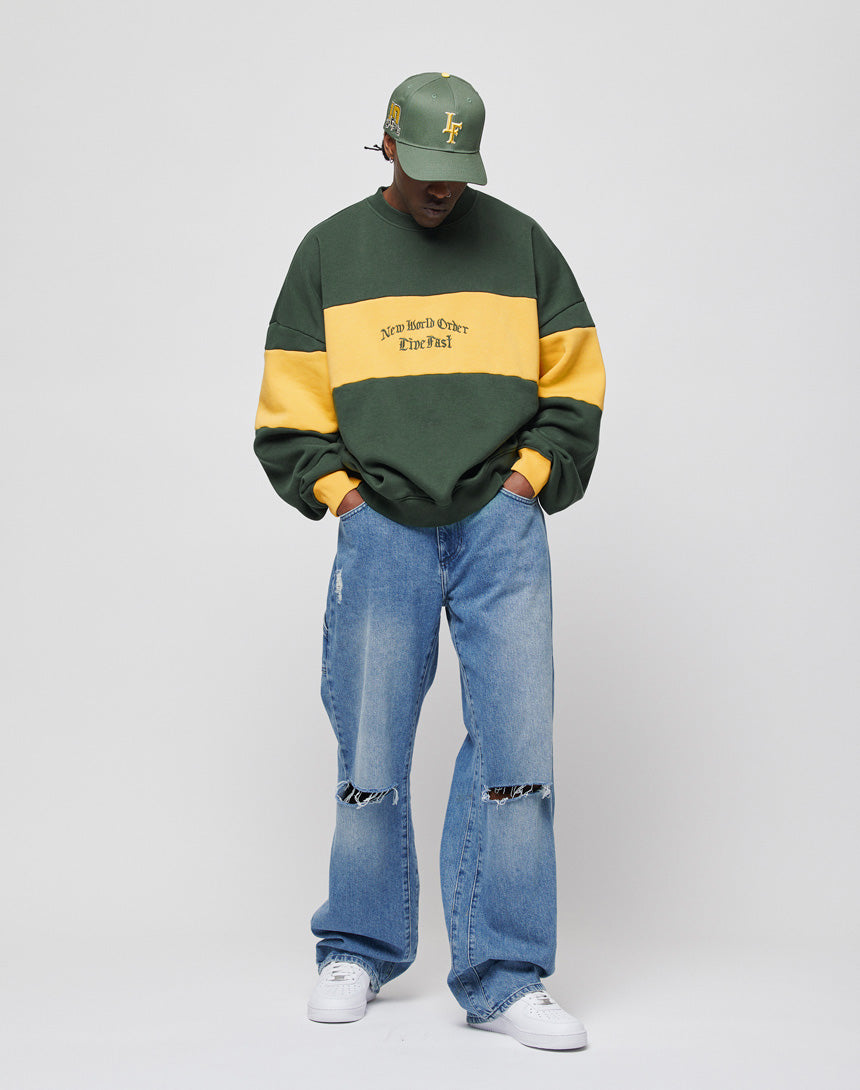 A person wears the LIVE FAST DIE YOUNG New World Sweater in green and yellow with loose ripped blue jeans, white sneakers, and a green cap, standing against a plain white background looking down with hands in the sweater pocket.