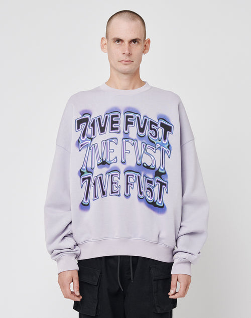 A person with a shaved head wears the LIVE FAST DIE YOUNG Triple Sweater—an oversized, light purple cotton sweatshirt with 7IVE FV5T printed three times in bold letters—paired with black pants against a plain white background.