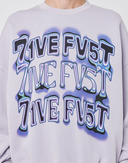 A person wears the LIVE FAST DIE YOUNG Triple Sweater, a light purple oversize-fit sweatshirt featuring 7IVE FV5T repeated three times in bold, distorted blue and black letters across the front for a cool vintage effect.
