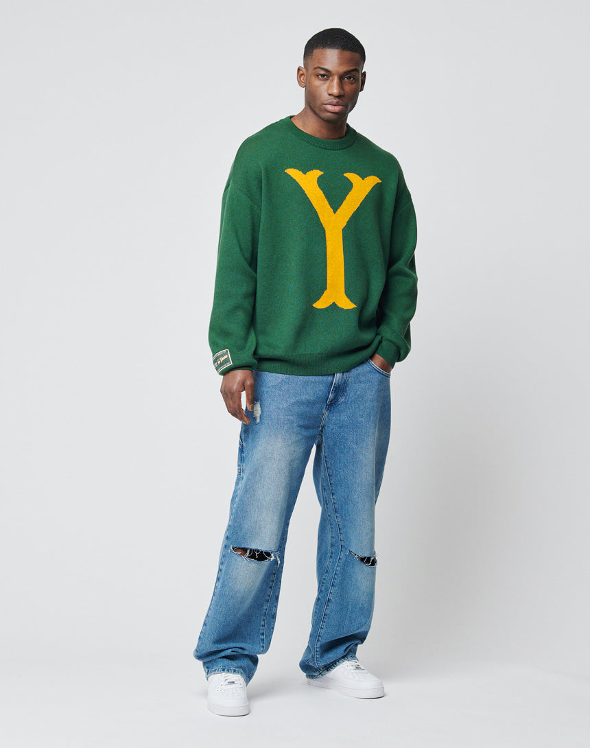 A man poses confidently with one hand in his pocket, wearing the LIVE FAST DIE YOUNG Y Knit Sweater featuring an oversized Intarsia Y logo, distressed blue jeans, and white sneakers against a plain backdrop.