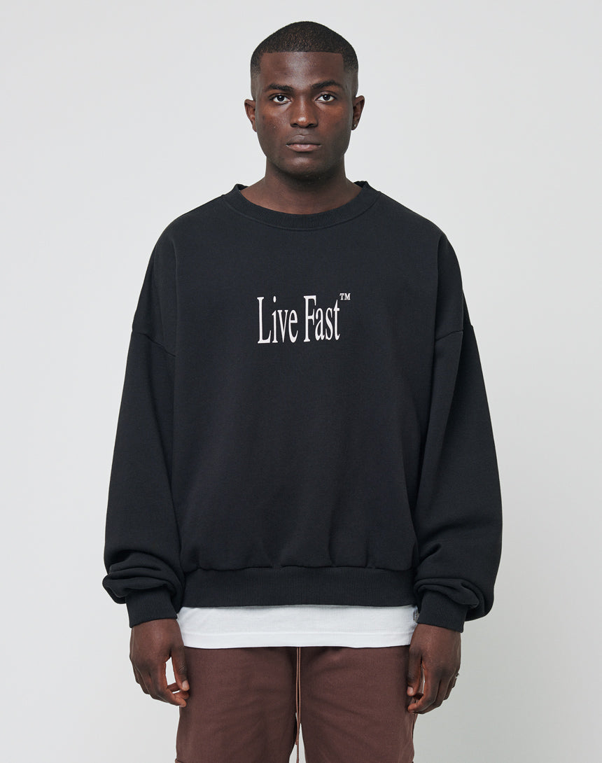 A man stands facing forward, wearing an LFDY Death Before Dishonor Sweater—black with white Live Fast™ text—over a white shirt and brown pants, set against a plain light background.