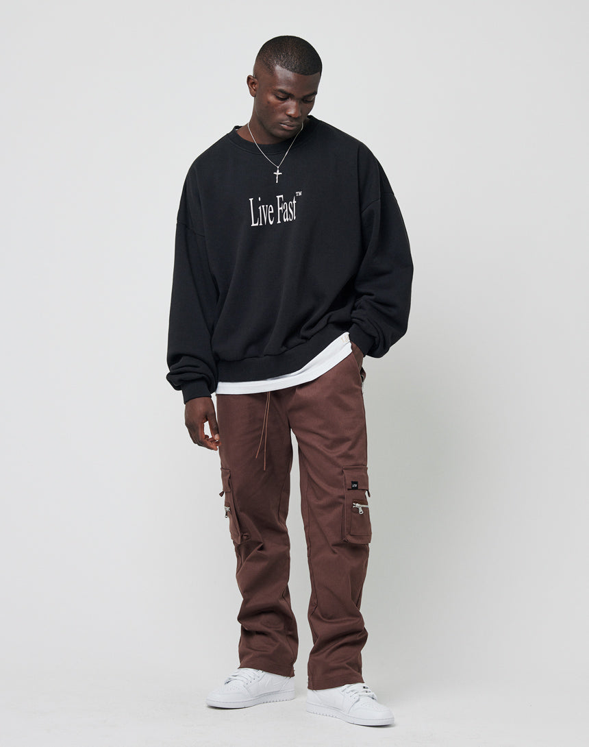 A man with short hair stands against a plain background wearing LFDY: the Death Before Dishonor Sweater in black over a white shirt, brown cargo pants, and white sneakers. He looks down, one hand in his pocket.