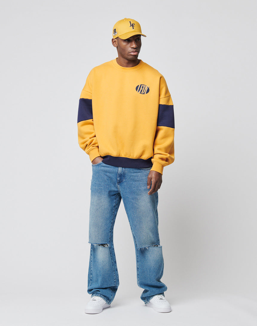 Wearing an LFDY® Sweater by LIVE FAST DIE YOUNG in yellow with navy sleeve stripes, paired with blue ripped jeans and white sneakers, a person stands against a plain light gray background.