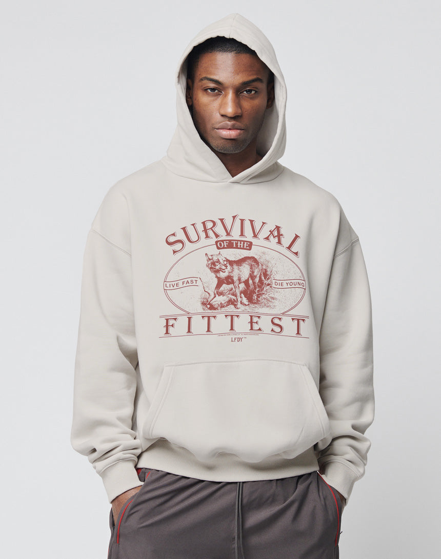 A man wears the LIVE FAST DIE YOUNG Survival Hooded, a light gray oversized cotton hoodie with a wolf graphic and Survival of the Fittest, Live Fast Die Young text. He stands against a plain background. Pflegeleicht and stylish.