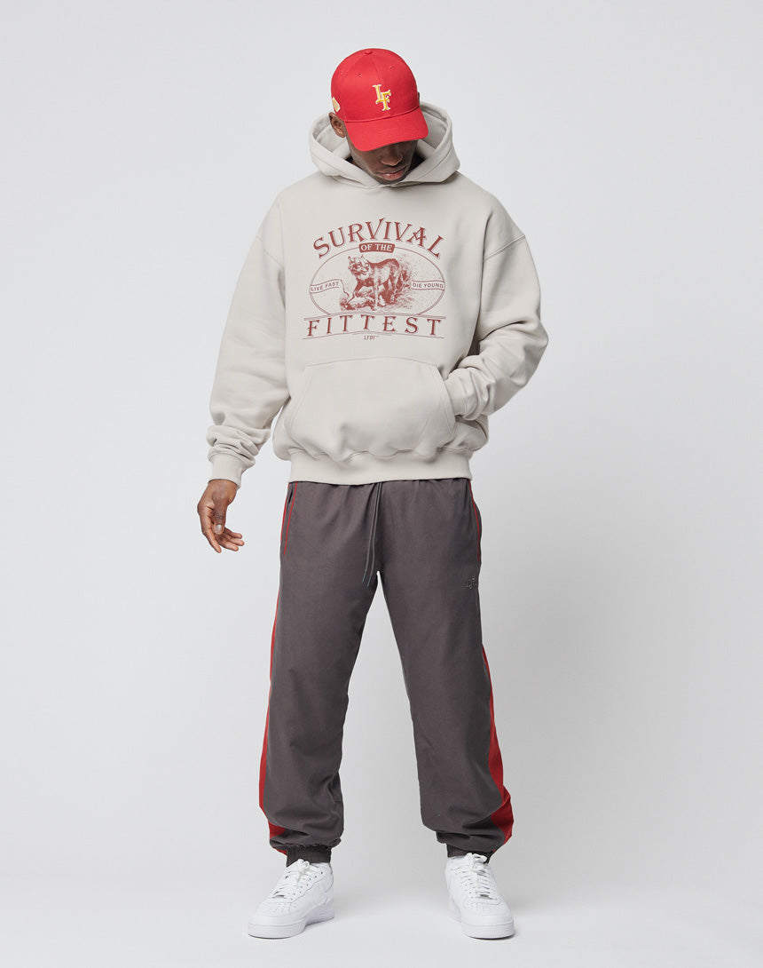 A person wearing a red cap, beige Regular Fit hoodie with “Survival of the Fittest” and Logo-Stick graphic, LFDY Trackpants by LIVE FAST DIE YOUNG in gray and red polyester, and white sneakers stands against a plain white background, looking down.