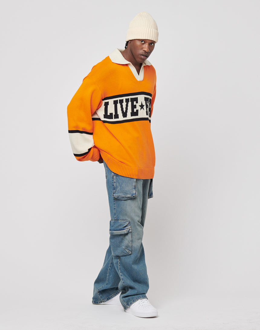A person in a cream beanie, loose blue cargo jeans, white sneakers, and a bright orange LFDY Knit Polo by LIVE FAST DIE YOUNG with "LIVE" and a star logo on the front stands against a plain white background.