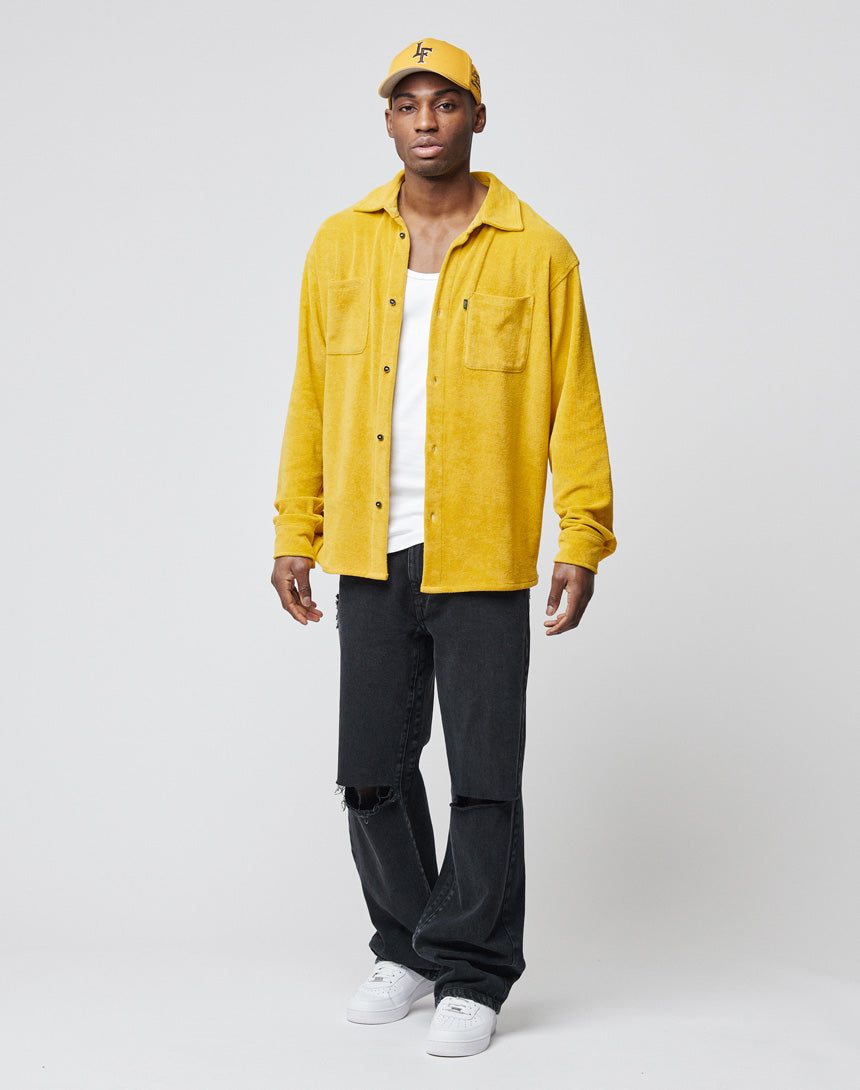 A man stands against a plain background, wearing the yellow LFDY Frottee Shirt by LIVE FAST DIE YOUNG in Oversize Fit over a white T-shirt, black ripped jeans, and white sneakers. The shirt is 100% Baumwolle. He faces forward with a neutral expression.