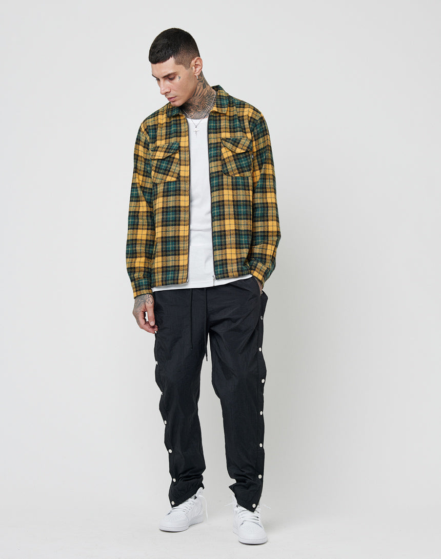 A tattooed person wears an oversize fit LFDY Zip Flannel in yellow and green plaid over a white T-shirt, with black snap button pants and white sneakers, standing against a plain white background and looking down.
