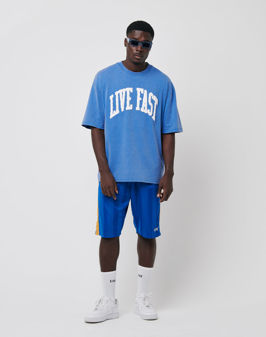 A man in sunglasses, a blue oversized LIVE FAST T-shirt, LFDY Taped Stripe Shorts with an Elastik-Bund and regular fit, white socks, and white sneakers stands against a plain white background.