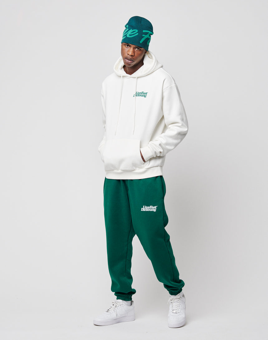 A person wears a LIVE FAST DIE YOUNG Basic Chest Hooded in white, green sweatpants, a matching green beanie, and white sneakers, standing hands-in-pockets against a plain light gray background.