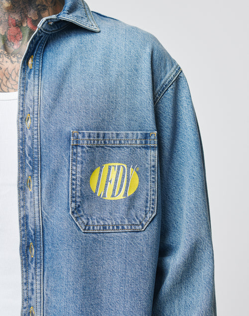 A person wears the LFDY® Denim Shirt by LIVE FAST DIE YOUNG, featuring a yellow and white LFDY logo on the chest pocket; part of their tattooed neck and a white undershirt are also visible.
