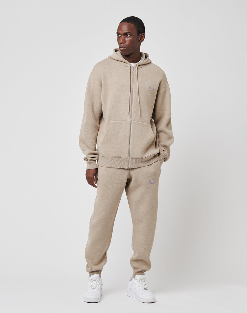 A man wears the LIVE FAST DIE YOUNG Knit Hooded Zip in beige with matching oversize joggers, featuring a drawstring hood (Kapuze mit Kordeln). He pairs the look with white sneakers and stands against a plain white background, left hand in pocket.