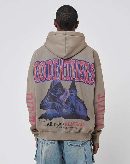 A person wears the LIVE FAST DIE YOUNG Godfathers Hooded in tan, an oversize fit with GODFATHERS in bold red and blue on the back, a vintage hooded figure by a church, āAll rights RESERVEDā text, and pairs it with blue jeans.