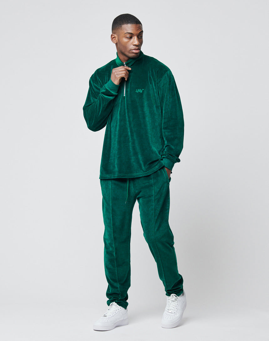 A man stands against a plain light grey background, wearing the LIVE FAST DIE YOUNG Velvet Halfzip in dark green velour with an oversize fit and metal zipper, paired with white sneakers. He looks left, holding the zipper near his neck.