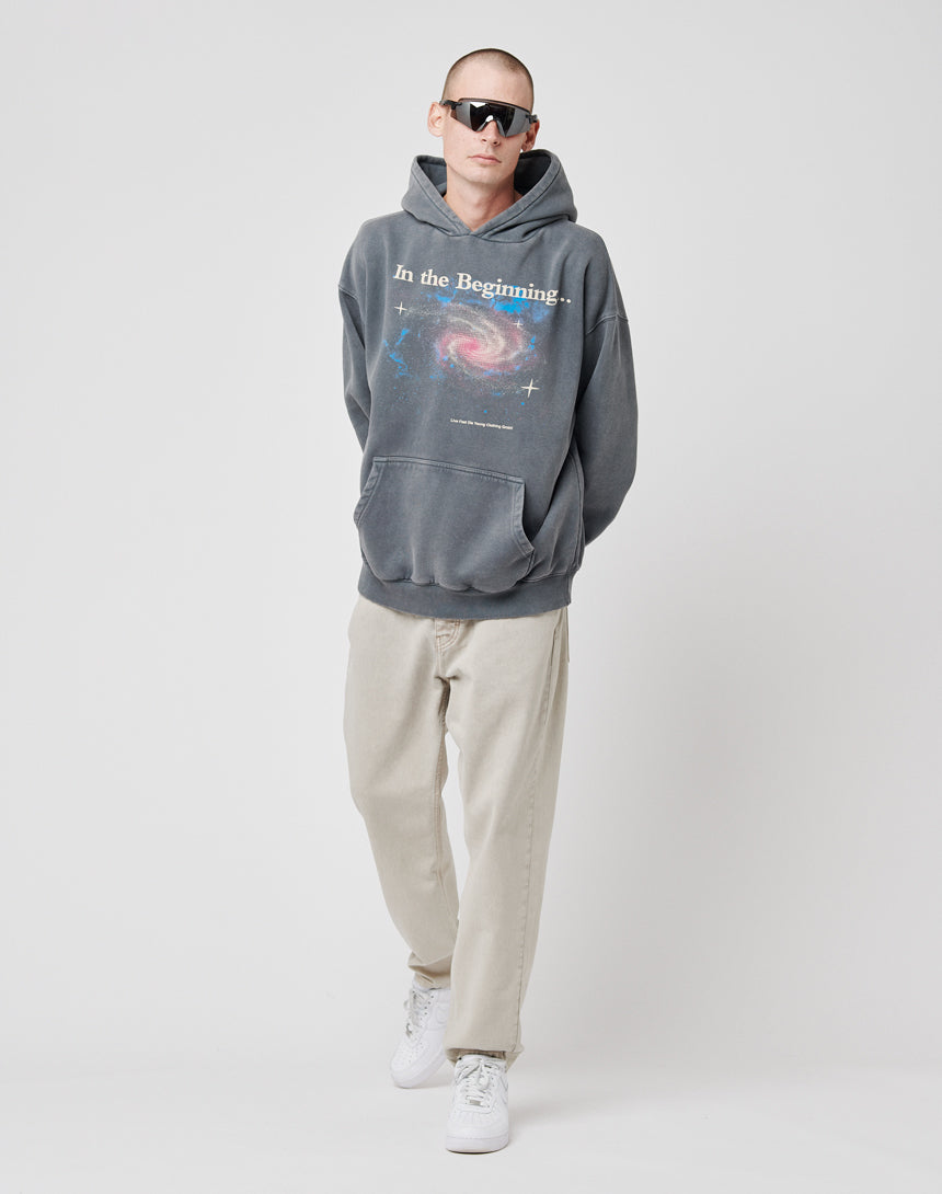 Wearing oversize sunglasses, light beige pants, and white sneakers, a person models the LIVE FAST DIE YOUNG Beginning Hooded—a gray hoodie with a vintage galaxy graphic and In the Beginning, blending Baumwolle Polyester for comfort.