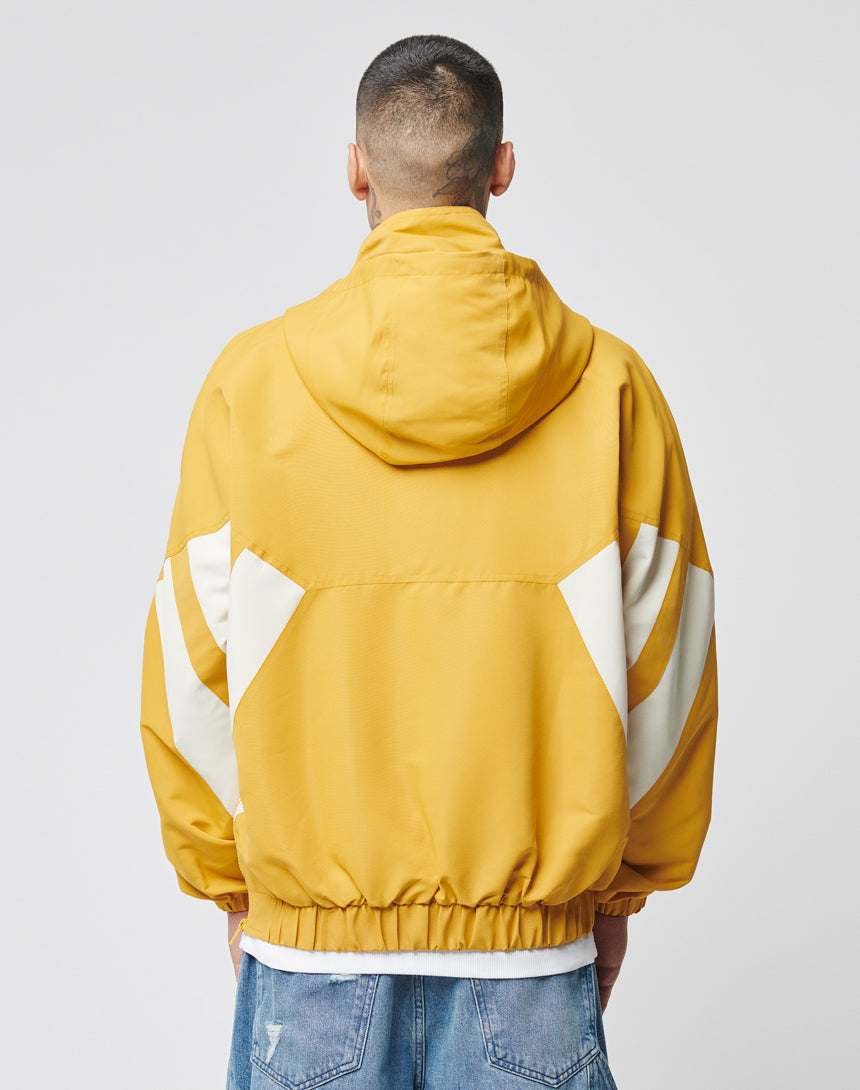 A person faces away from the camera wearing a yellow LIVE FAST DIE YOUNG Half Zip Hooded Jacket with white geometric sleeve and back designs, paired with blue distressed jeans.