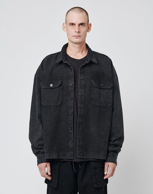 A man with a shaved head wears the LIVE FAST DIE YOUNG Vintage Dyed Flannel, a loose-fitting black button-up in 100% cotton with an oversize fit and two front pockets, standing against a plain light gray background.