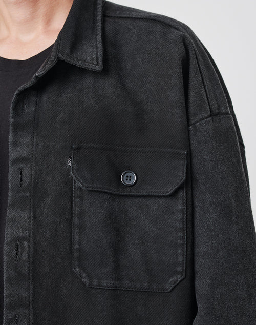 Close-up of a person wearing the LIVE FAST DIE YOUNG Vintage Dyed Flannel, an oversize fit black button-up shirt with a chest pocket and black buttons, layered over a black t-shirt. Only the lower face and upper torso are visible.