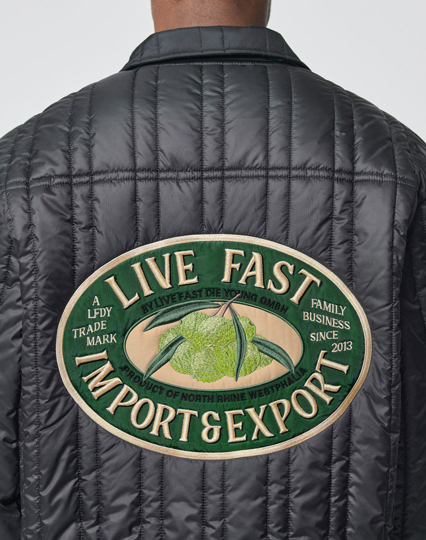 Someone wears the Import & Export Jacket by LIVE FAST DIE YOUNG—an oversize black jacket with a large back patch reading “Live Fast Import & Export,” plus a green fruit graphic and text about a family business founded in 2013.