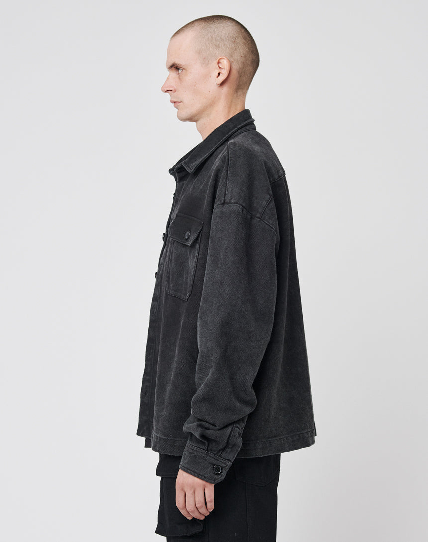 A person with a shaved head stands in profile facing left, wearing the LIVE FAST DIE YOUNG Vintage Dyed Flannel, an oversized black button-up jacket with chest pockets, and black pants against a plain white background.
