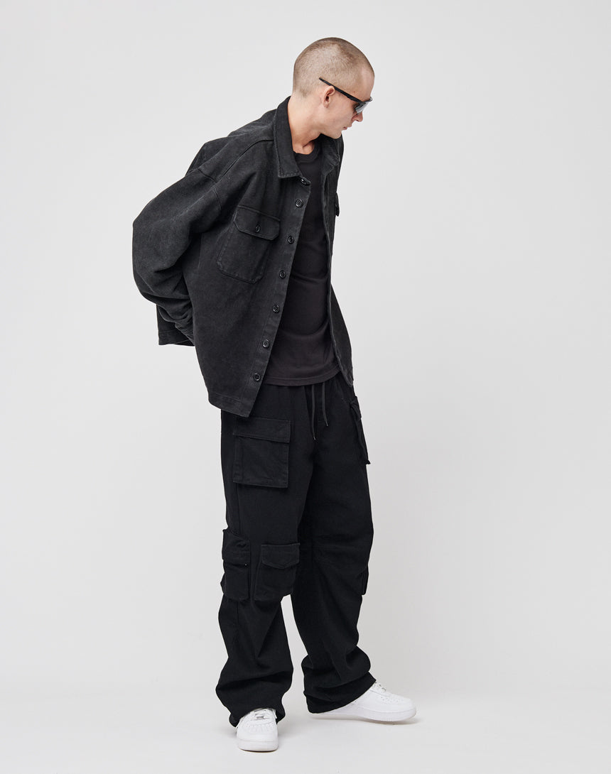 A person in LIVE FAST DIE YOUNG’s Vintage Dyed Flannel overshirt (100% Baumwolle), sunglasses, black cargo pants, and white sneakers stands against a white background, gazing to the side with hands behind their back.