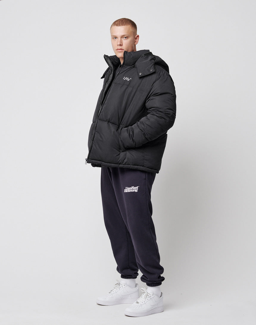 A person stands against a plain white background wearing the LIVE FAST DIE YOUNG Basic Winter Jacket in an oversized fit, dark sweatpants, and white sneakers, looking slightly to the side with a neutral expression.