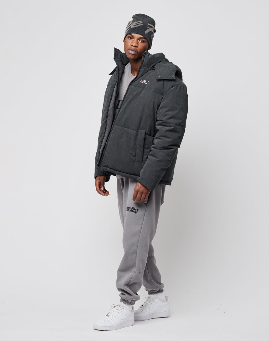 A man in a LIVE FAST DIE YOUNG Basic Winter Jacket, gray sweatpants, gray beanie, and white sneakers stands against a plain light background, gazing slightly to the side with a relaxed posture.