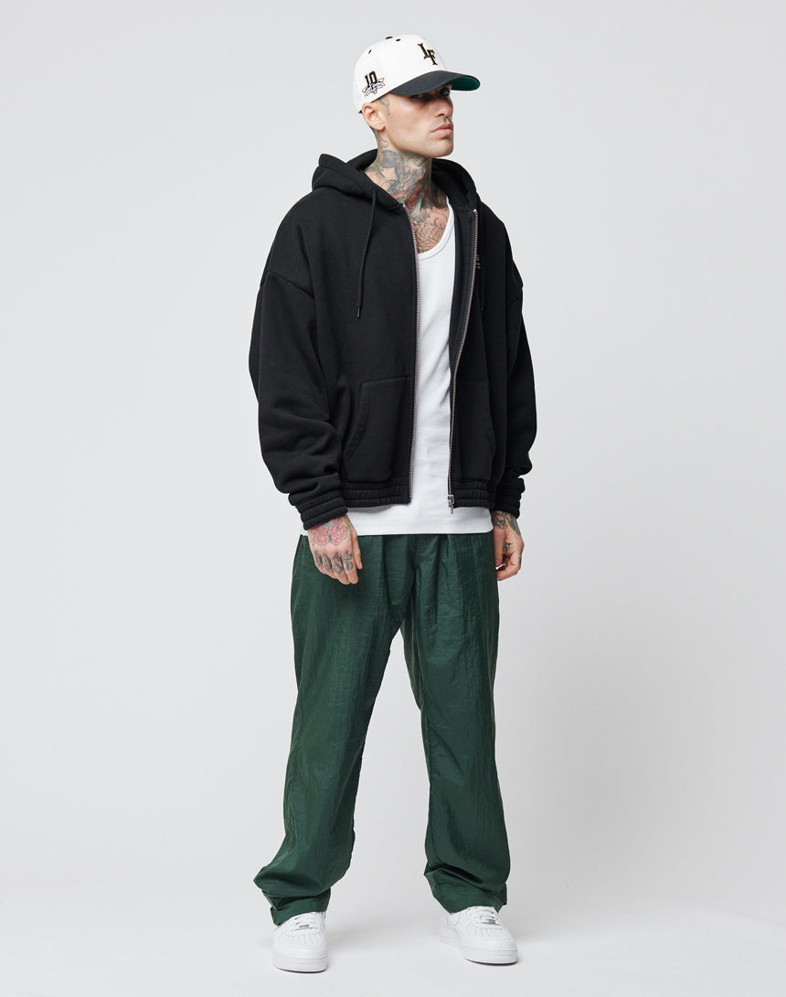 A man in a black zip-up hoodie over a white tank top, LIVE FAST DIE YOUNG Nylon Jogger pants with Breiter Elastikbund, white sneakers, and a white cap stands against a light gray background, showing tattoos on his neck and hands.