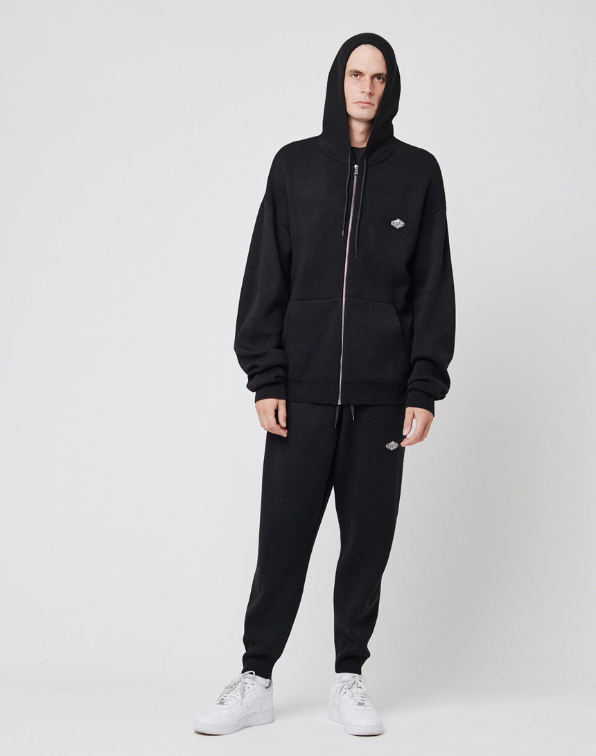 A person wearing an oversize fit black LIVE FAST DIE YOUNG Knit Hooded Zip with the hood up, matching black sweatpants, and white sneakers stands against a plain white background.