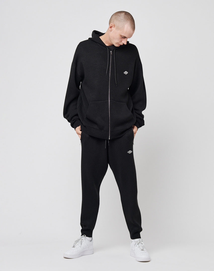 A person wears LIVE FAST DIE YOUNGs Knit Jogger and black zip-up hoodie with a logo patch, both featuring an elastic waistband, standing hands-in-pockets against a plain background with white sneakers.