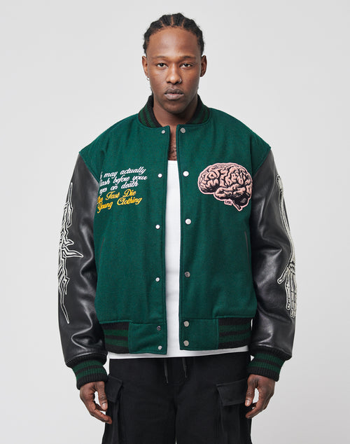 A person stands facing forward wearing the Life College Jacket by LIVE FAST DIE YOUNG—green with leather sleeves, logo snap buttons, embroidered brain and text on the chest, and white graphics on the sleeves—over a white shirt and black pants.