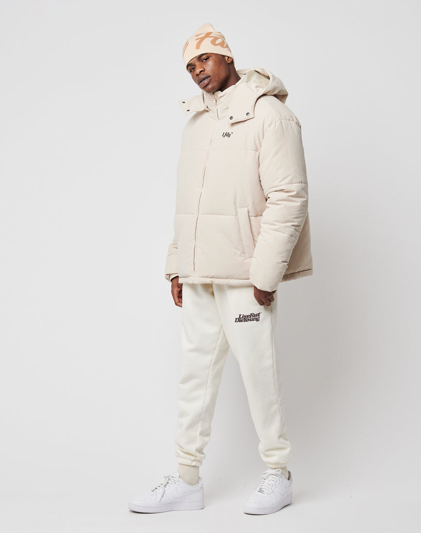 A man stands against a white background wearing an oversize beige LFDY Basic Winter Jacket, matching beanie, white logo sweatpants, and white sneakers. The padded jacket is wind- and weatherproof for style and comfort. He poses relaxed with one hand in his pocket.