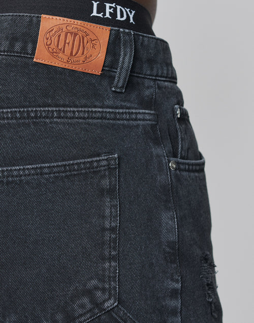Close-up of a person wearing LIVE FAST DIE YOUNG Oversize Ripped Denim jeans with an oversize fit, straight leg, brown leather LFDY waistband label, slight distressing, back pocket, and LFDY-branded black underwear visible above.