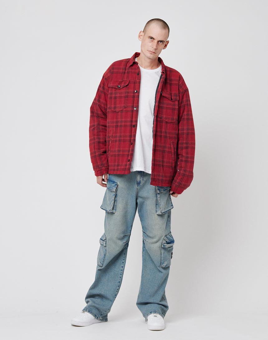 A person with a shaved head stands against a white background, wearing a LIVE FAST DIE YOUNG Padded Flannel Jacket over a white T-shirt, baggy blue cargo jeans in Oversize Fit Body, and white sneakers. Their expression is neutral.