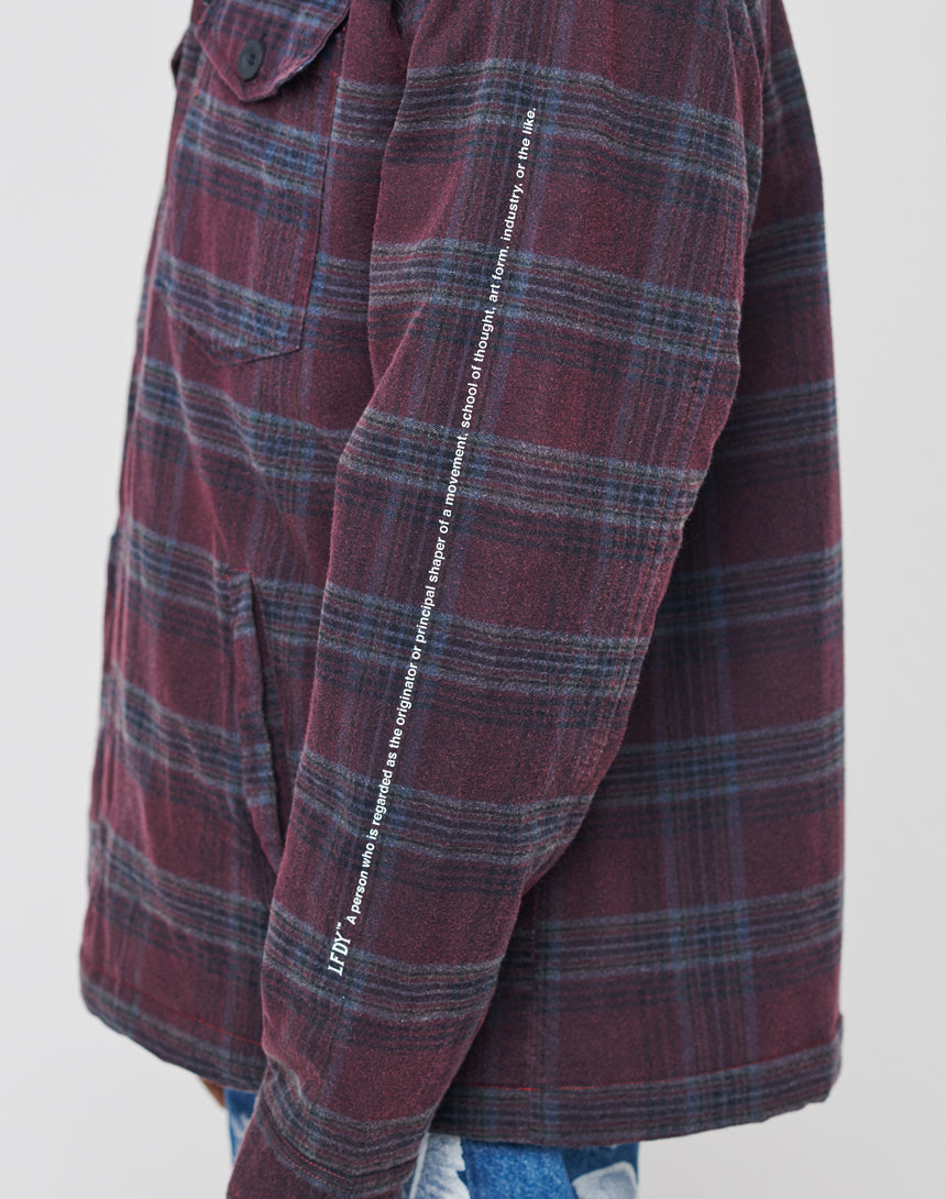 A close-up shows a persons arm in the LIVE FAST DIE YOUNG Padded Flannel Jacket—maroon and navy plaid with a vintage effect, oversize fit, chest pocket, and white vertical text on the sleeve—styled with blue jeans.