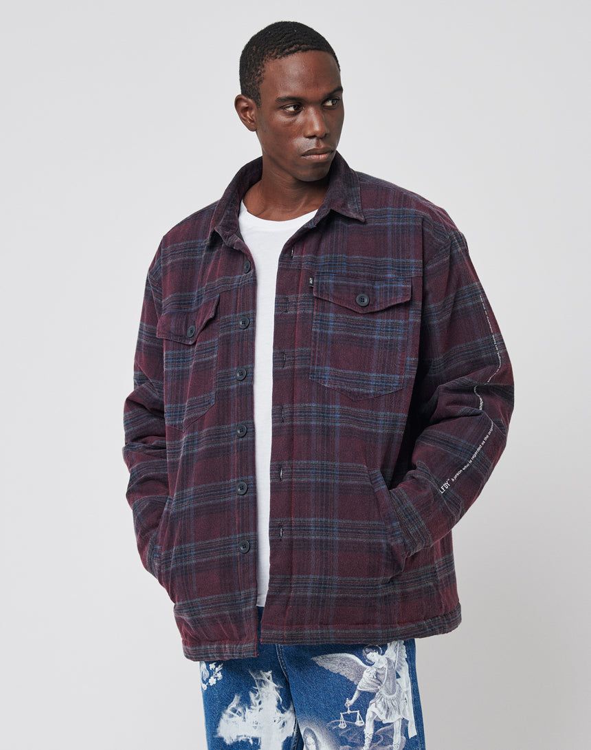 A man wears a LIVE FAST DIE YOUNG Padded Flannel Jacket with a maroon and navy plaid vintage effect over a white shirt, pairing it with blue jeans featuring white printed artwork, standing against a plain light background.