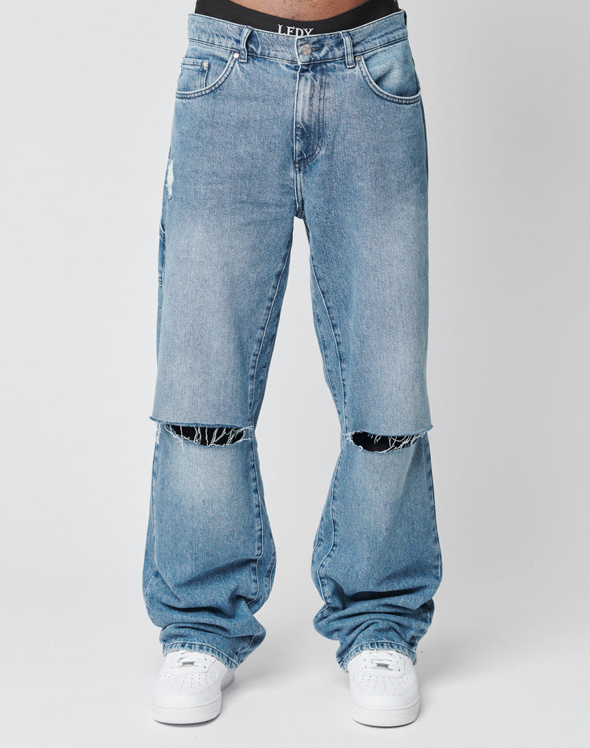 A person wears LIVE FAST DIE YOUNG Oversize Ripped Denim—light blue, oversized fit, large knee slits, straight leg—paired with white sneakers and a visible waistband, set against a plain background.