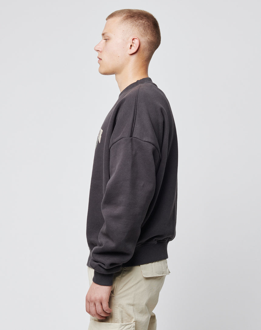 A young man with short hair stands in profile against a plain light background, wearing the LIVE FAST DIE YOUNG Live Fast Die Young Chest Sweater and light-colored cargo pants, his arms relaxed at his sides.