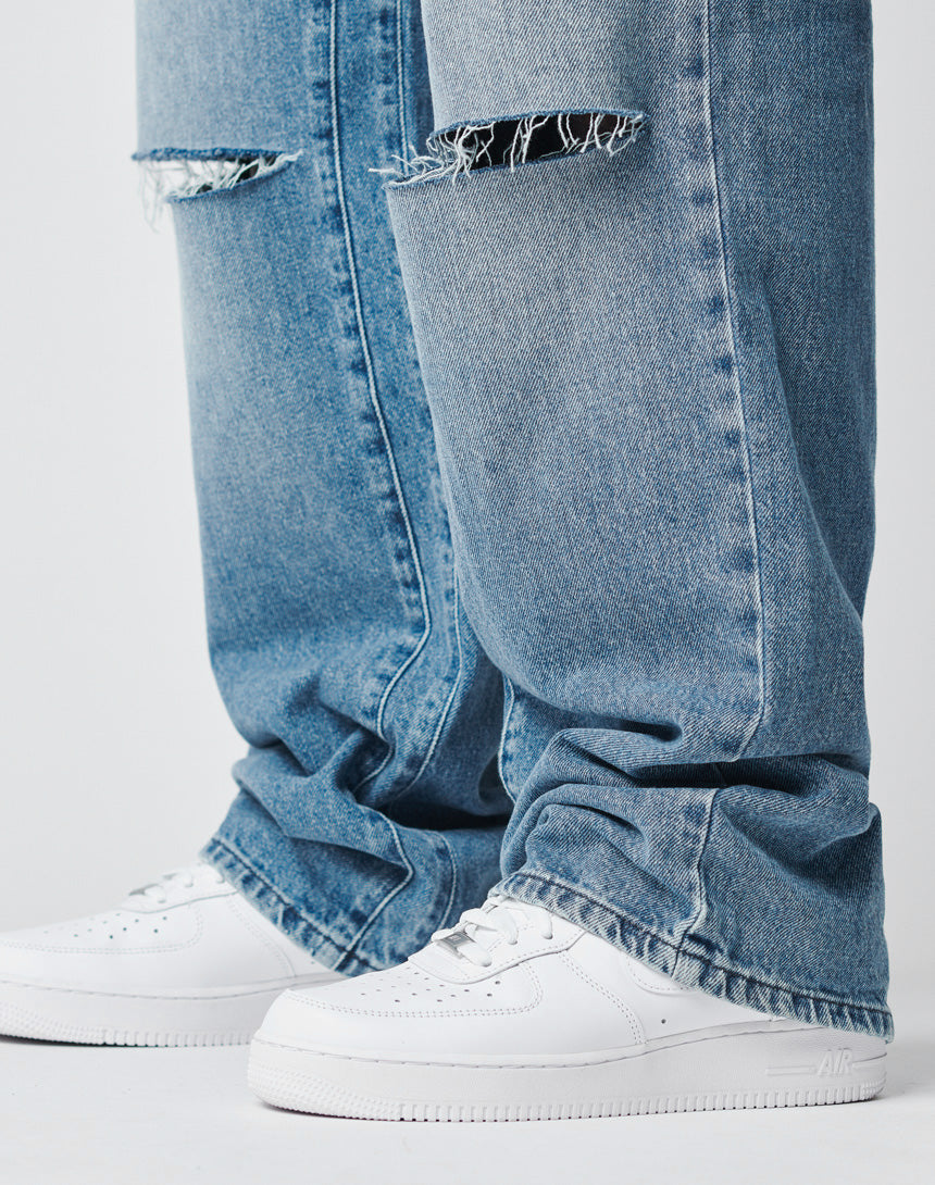 A close-up of a person wearing LIVE FAST DIE YOUNG Oversize Ripped Denim, light blue with knee slits and straight leg, paired with white Nike Air Force 1 sneakers, standing on a white surface.