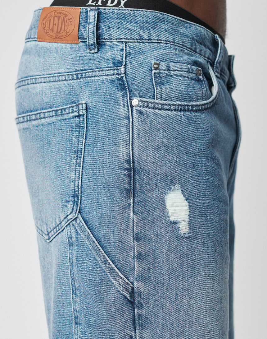 Close-up of a person wearing LIVE FAST DIE YOUNG Oversize Ripped Denim in light blue, featuring a small thigh rip and leather brand patch, with a visible black waistband with white text.