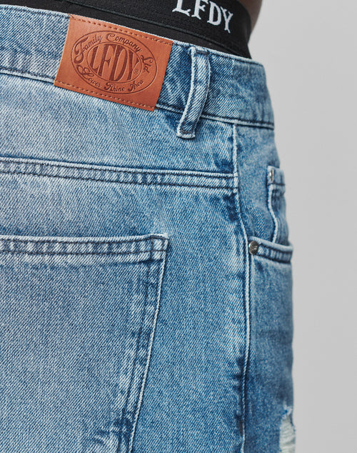 Close-up of Oversize Ripped Denim by LIVE FAST DIE YOUNG, showing blue denim with a brown leather brand patch above the back pocket and part of a black waistband with LFDY text at the top.